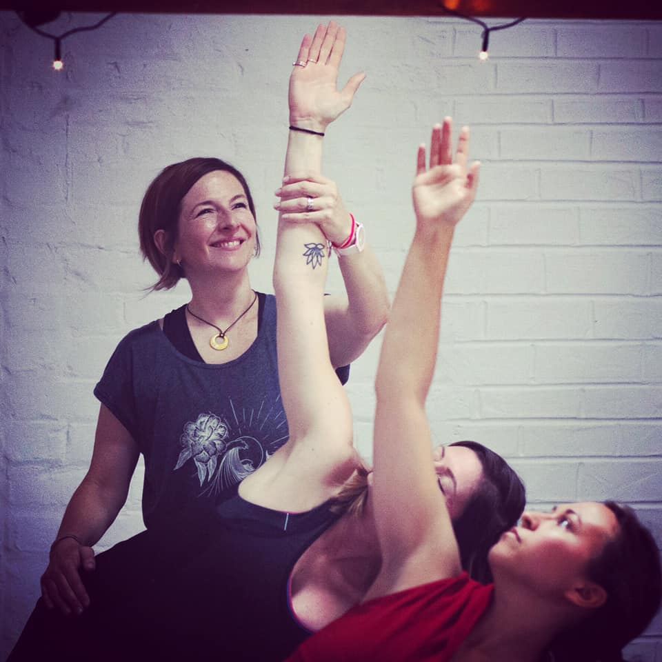 A teacher smiling as she guides a student's raised arms during a Hatha Yoga Teacher Training session