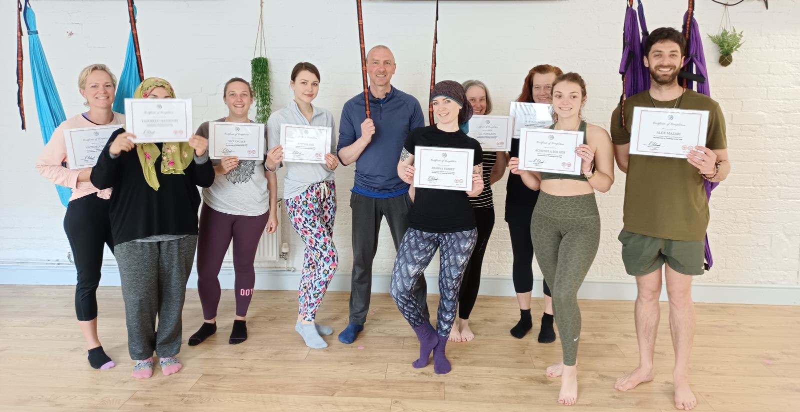 A group of Aerial Yoga teacher training graduates holding their certificates beneath the aerial swings