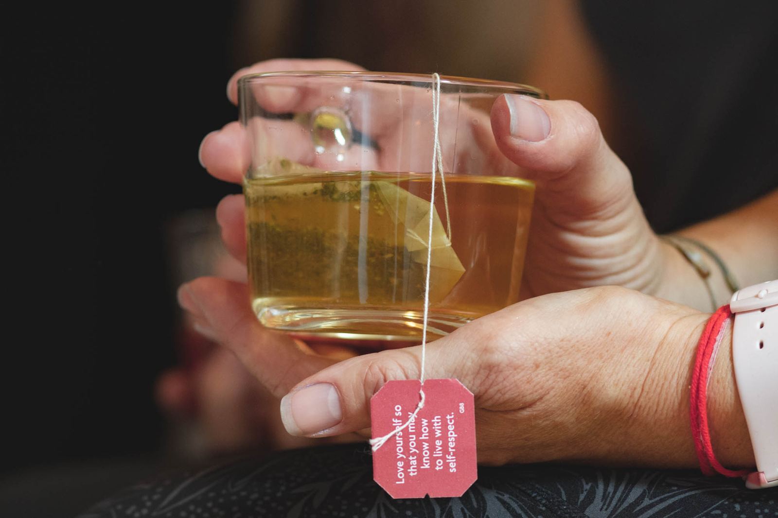 Hands holding a glass of herbal tea with the tea bag tag hanging down