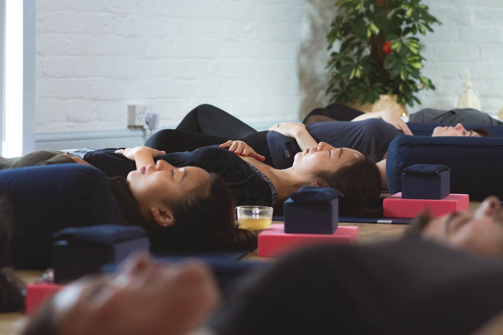 Students lying on their backs on mats in a quiet studio, resting with bolsters and blocks around them