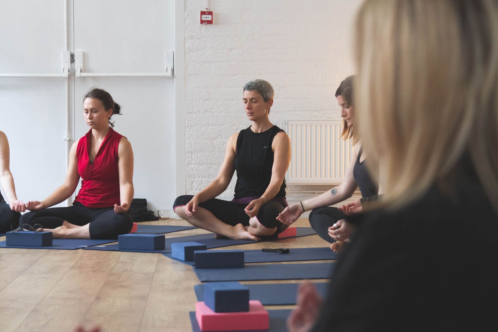 Students sitting cross-legged on their mats with eyes closed, hands resting on their knees, yoga blocks in front of them