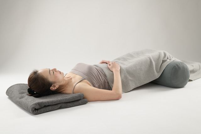 A woman resting in a supported Restorative Yoga pose with a bolster under her knees and a blanket