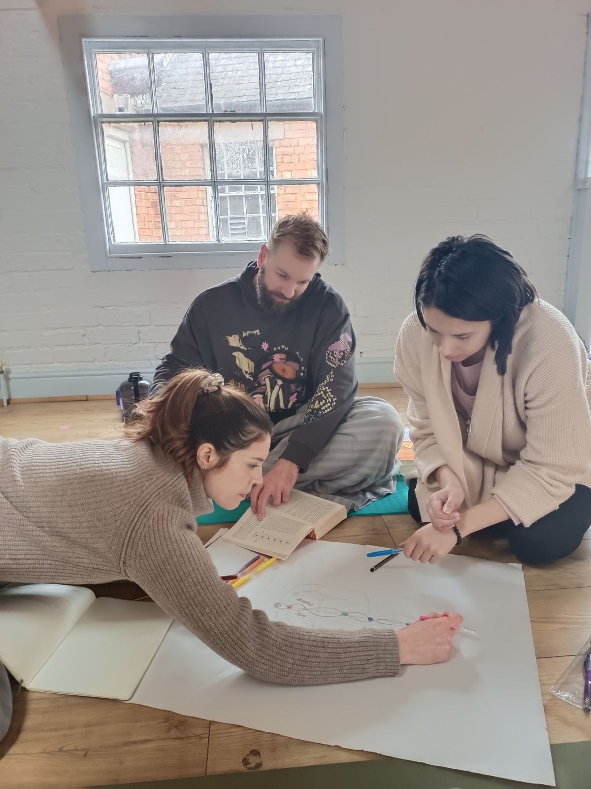 Three trainees sitting on the studio floor, drawing and studying together on a large sheet of paper