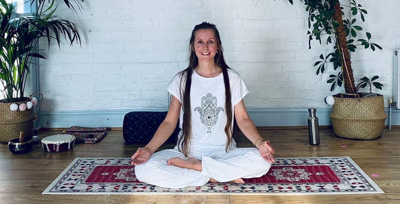 A smiling Kundalini teacher dressed in white, sitting cross-legged on a patterned rug with a drum and singing bowl nearby