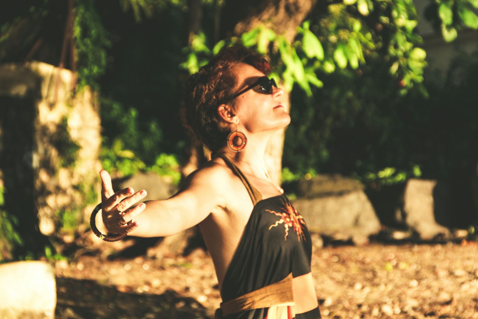 A woman standing outdoors in dappled sunlight, arms open wide and face turned to the sky