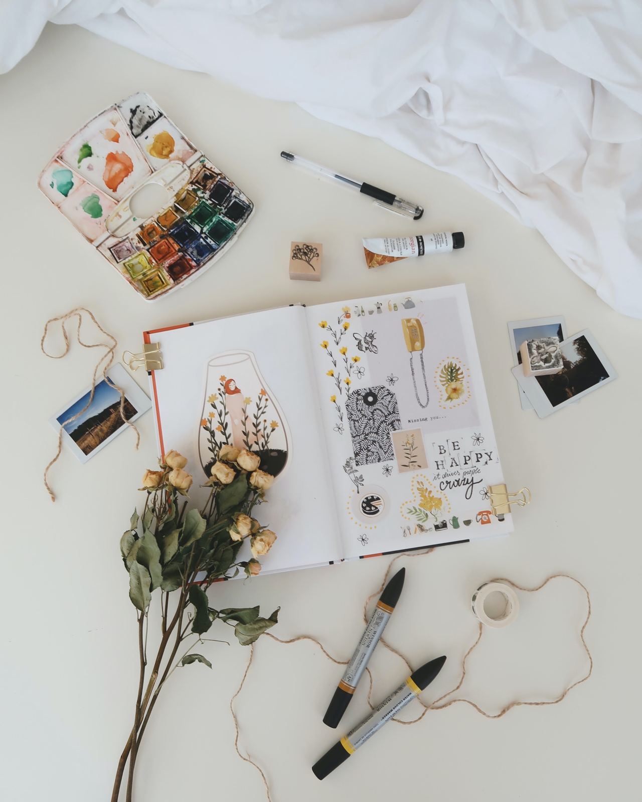 An open art journal with watercolours, pens, photos and dried flowers