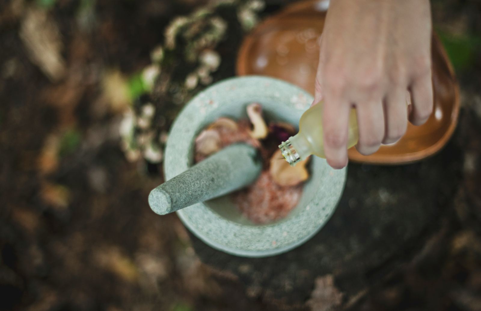 A hand pouring oil into a stone pestle and mortar filled with salt and dried petals
