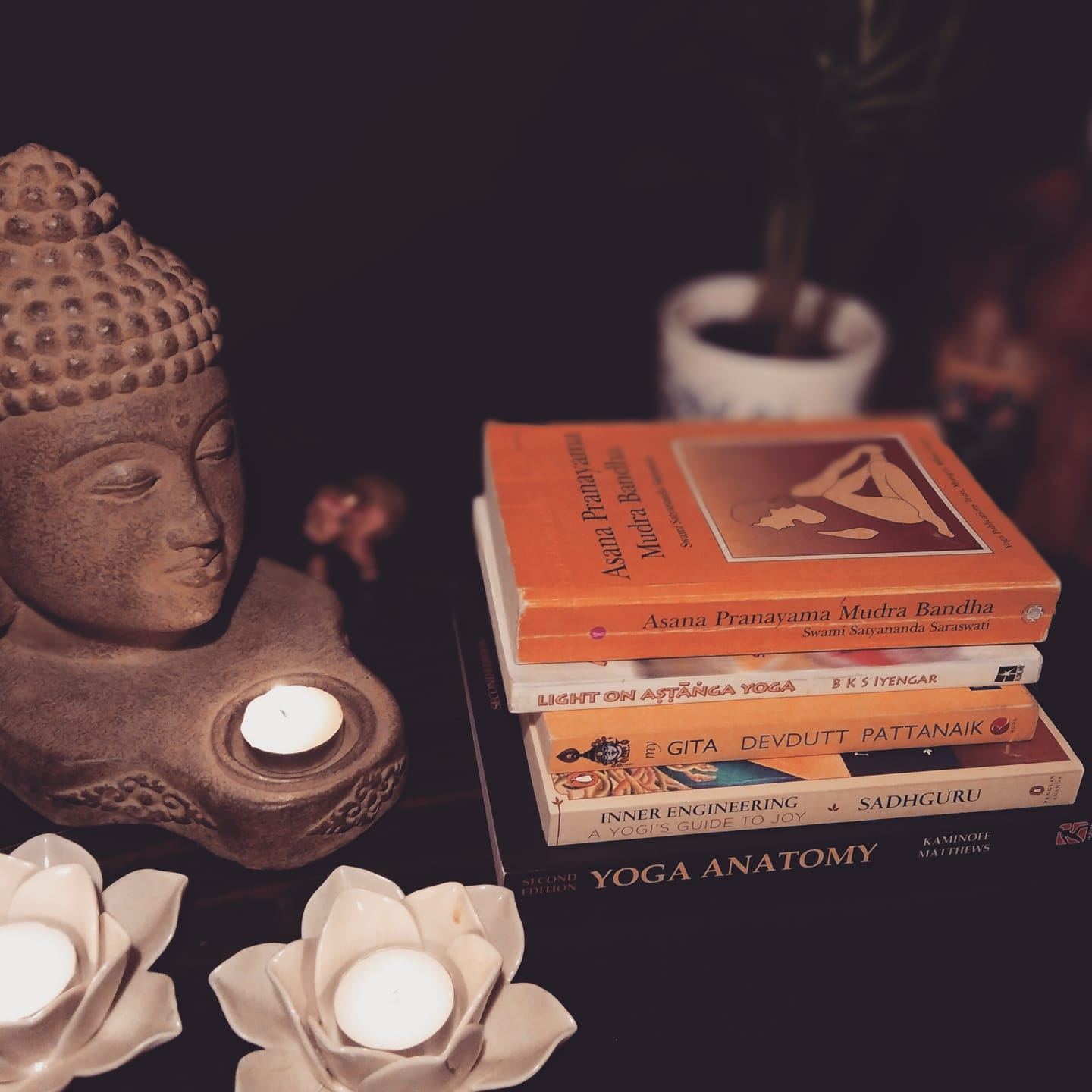 A stack of yoga books, including Light on Ashtanga Yoga and Yoga Anatomy, beside a Buddha head and candles