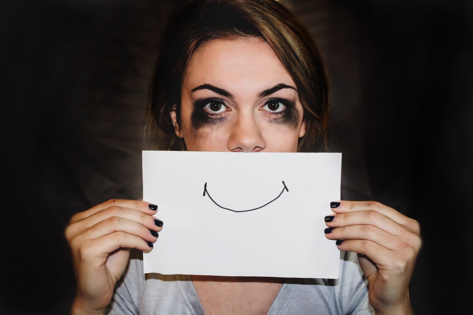 A woman with tear-smudged eye make-up holding a drawn smile in front of her mouth