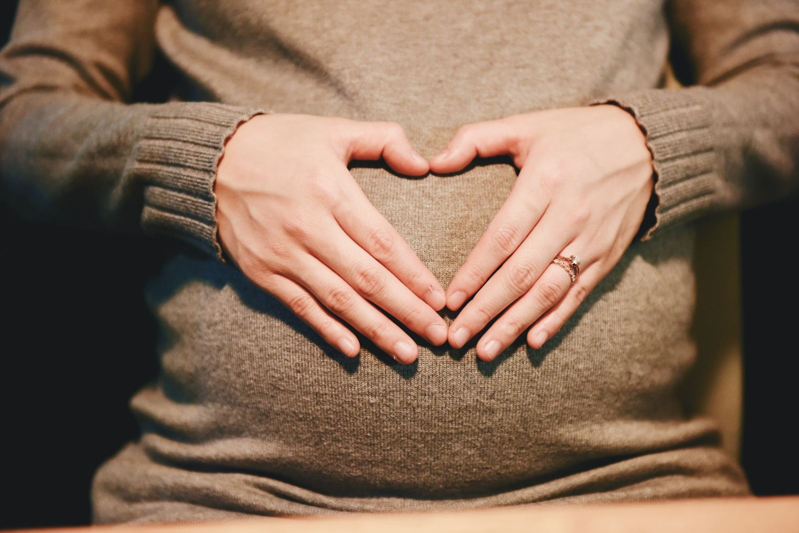 A pregnant woman in a soft jumper resting her hands in a heart shape on her bump