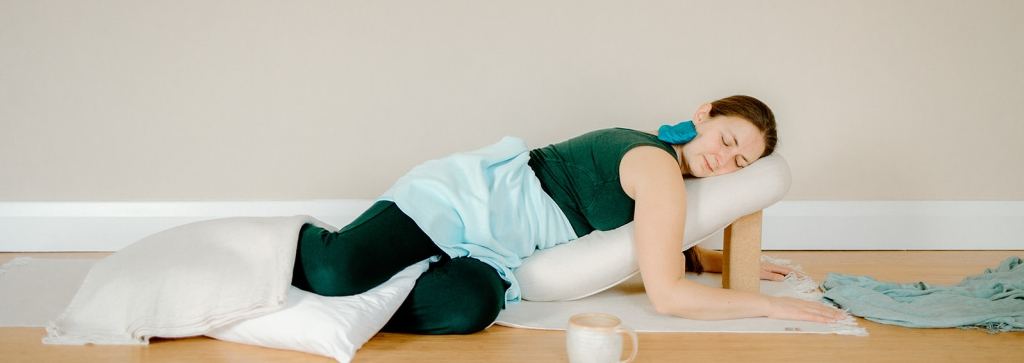 A woman resting face down over a bolster propped on a block, eyes closed, with a blanket over her legs