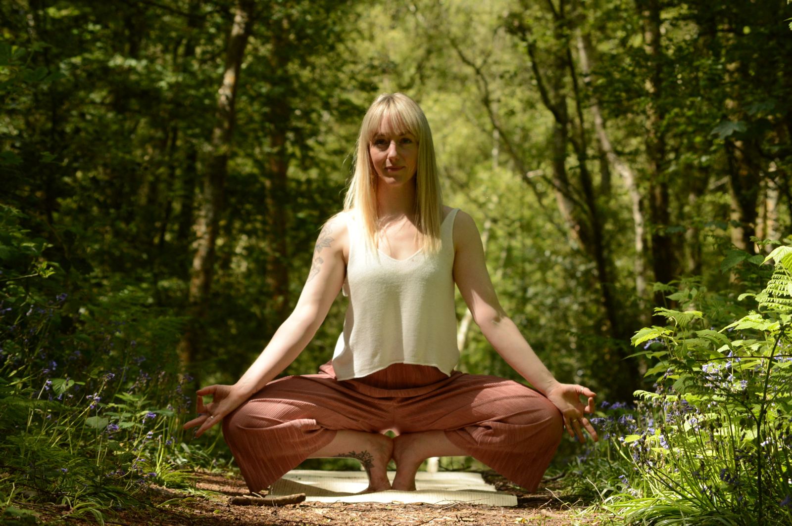 A yoga teacher in a deep squat with hands in mudra, in sunlit bluebell woods